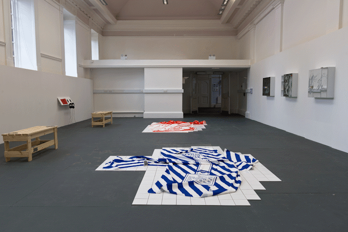 5 wall mounted cabinets, two floor sculptures, a TV, two benches, a shelf with headphones and publications are laid across a room