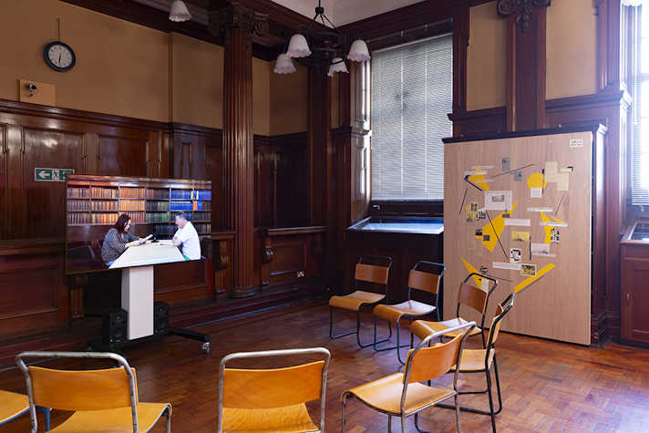A digital screen surrounded by chairs in a semi circle. Leaning against the wall is a large piece of MDF with images and writing on it.