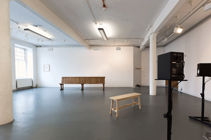 A pew, a bench, speakers on stands in a gallery