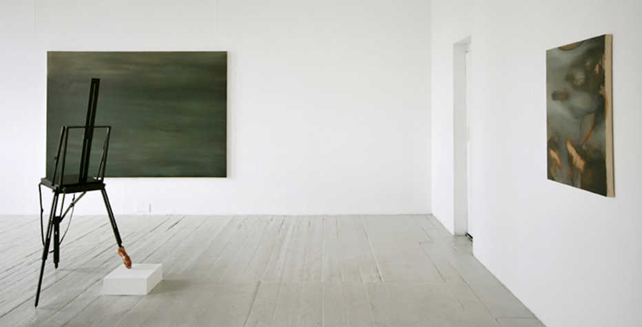 two dark grey green blurred paintings on adjacent walls. in the middle of the room is a black easel,