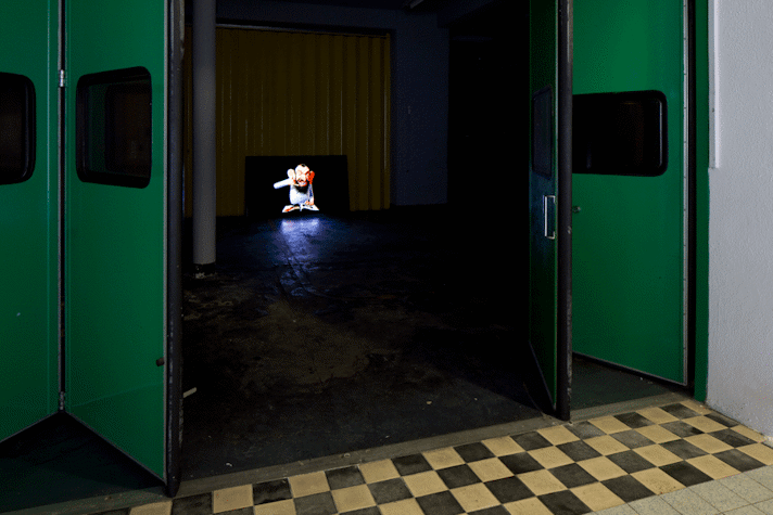 There are folding, sliding doors which are half open. They lead to a dark room with a monitor at the end of the room. The Screen of the monitor has the image of a puppet man. He has big ears, big nose and a beard. He is reading a book, while looking at something and making the gesture insinuating asking someone to call him.