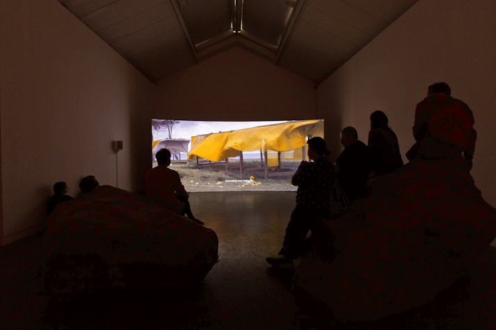 Installation view, people viewing film screen showing large orange fabric blowing on a structure