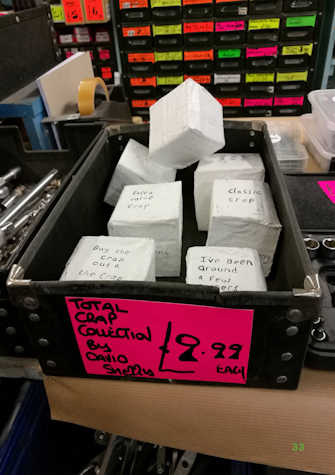A black plastic box near other boxes containing tools. This box contains plaster boxes that say "classic crap", "extra value crap", "buy the crap out a the crap", etcetera. The box is labeled with a pink sign, with black marker that reads "Total crap collection by David Sherry. £9.99 each"