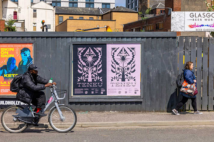 A two posters are displayed outside on a dark wooden fence, people walk past and there is a person on a bike.