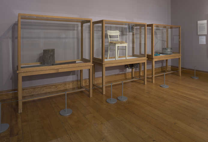 3 wood and glass display cases. in the first there is a tree stump, the second a chair and the third a metal cylinder