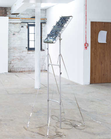 Free standing metal sculpture placed in an white room with white pillars. The sculpture has a thin tubular frame, with a panel of weaved thin sheet metal.