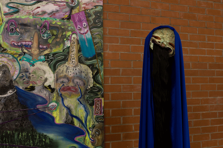 Detail of an artwork. There is a section of a painting on paper, hanging on a red brick wall. There are surreal, psychedelic faces merging with and emerging out of a landscape. There is the edge of an inky lake below a mountain, a crying face whose tears lead a river into another face's mouth. Next to the painting a mask with half closed eyes and a wide mouth is adorned with a cape of blue fabric, dark brown hair falls from its mouth.