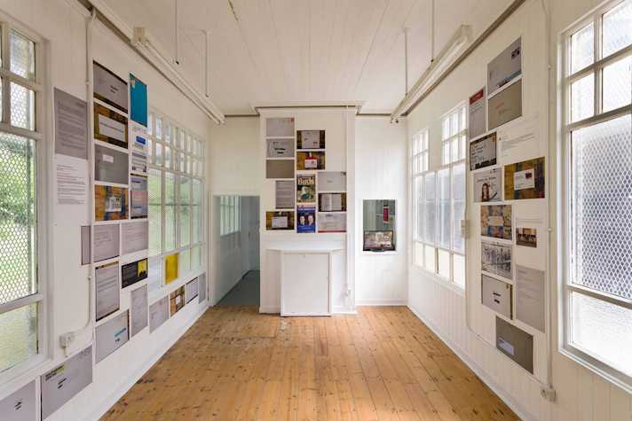 colourful photocopied sheets are pasted to the walls of a small gallery space. The sheets show letters, emails documents and other text-based media.