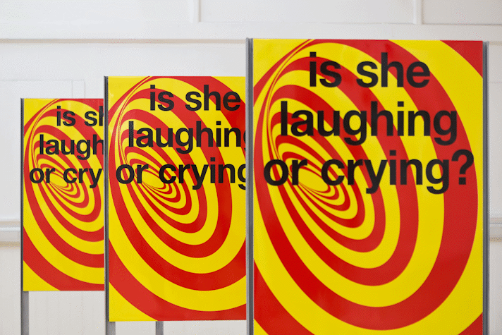 Three identical metal stands hold three identical portrait oriented graphic posters with a red and yellow swirl and black text overlaid. The text reads 'Is she laughing or crying?'.