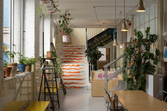 Room with natural light coming from a window to the left and filled with plant pots. Stairs at the end with carrot drawings go up and it reads "glazing workshop". There are wooden benches and tables.