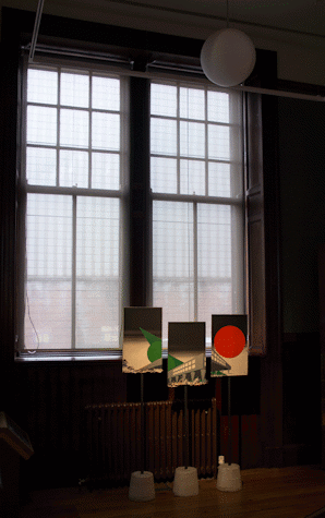 From a further away perspective and seeing it from the side we see a dark room, with windows with light behind and a radiator below, we see three rectangular painitngs that make up one in different altitudes. There is a grey building with a lot of windows and a green pointy shape to the left and a red circle to the right.