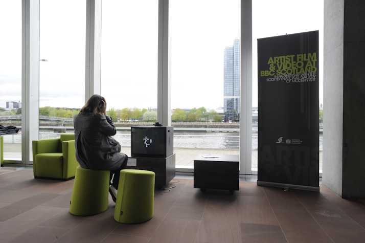 person sat on a green stool watching a crt monitor. there is a a sign reading: artist film & video at BBC Scotland in partnership with the Scottish national gallery of art