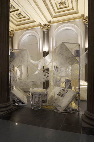 A metal installation of web-like plastic materials and other electronic and artwork is illuminated with white light in front of an elegran wallas part of an art installation in the Gallery of Modern Art, Glasgow International 2018.