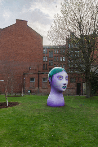 Profile left view of a big, tree-sized bust of a woman with purple skin, green eyes, thin eyebrows and green hair and lips. It is positioned in a park.