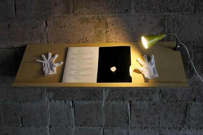 book on a shelf attached to a wall. there is a lamp clipped to the edge of the shelf. on both sides of the book there is a white cotton archival glove.