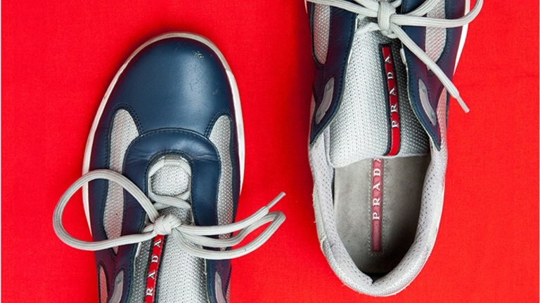 A photograph of a pair of blue and gray trainers with a red prada logo on the tongue. They are placed on a bright red surface,