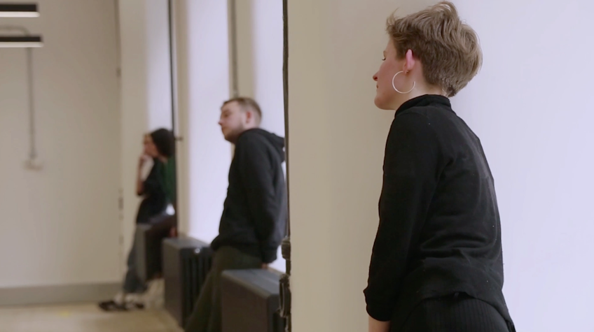 Three people dressed in black are leaning towards the window or wall on the right.