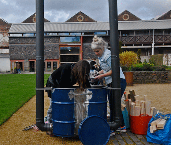 Two women are in the process of putting garbage into a couple of joint trash cans. One of them is reaching their hands into the cans, while the other is waiting to put dry waste into it. Two pipes are connected to the lower sides of either sides of the trash cans. On the left, there are packets and buckets of wooden planks. There is a building in the background.