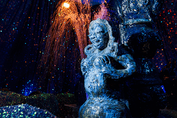 A ceramic figure of a nude woman with water spraying behind, surrounded in blue sparkly material.