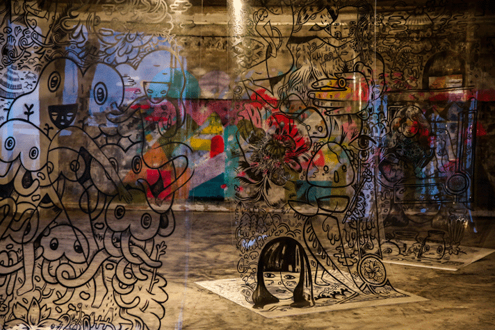 A glass wall has black drawings and you can see behind a wall of concrete with colorful grafitti and painting.