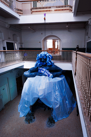 A large tented figure is displayed in a hall. It is wearing a long blue dress and their hands are rested on their hips. In the centre of their skirt there is a parting where people can enter.