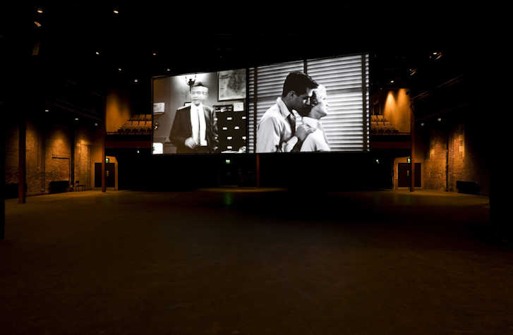 two projection screens hanging side by side in tramway 1. on the left is a man standing in a suit. on the right is a person stood behind another person with their hands on their shoulders in embrace.