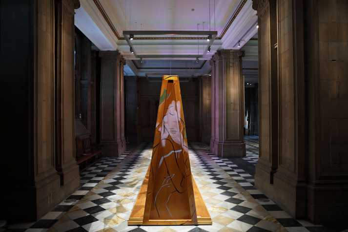 A stained wood structure sits on a checkerboard marble floor in between stone columns. The structure is painted with fast gestural strokes of paint depicting a nude figure looking over their shoulder