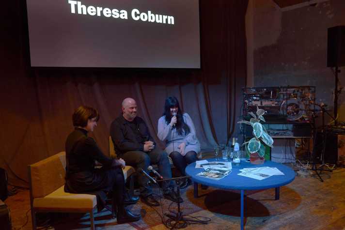 In a dim-lighted room Theresa Coburn and Stephen Sutcliffe in conversation with Claire Biddle with a proyector screen on top of them that reads "Theresa Coburn"