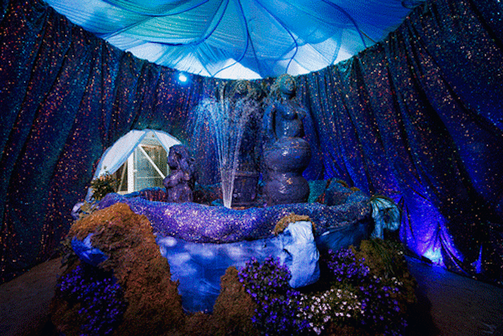 A blue water fountain featuring ceramic nude figures is underneath a domed blue ceiling with walls decorated by sparkly blue fabric.