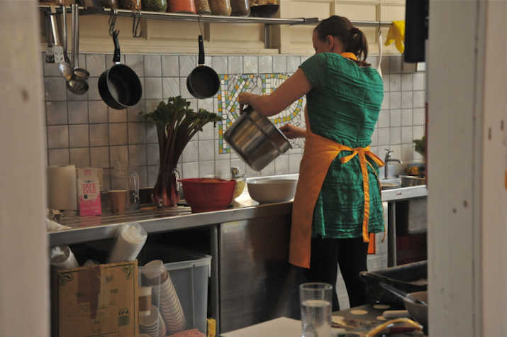 In a kitchen there is a woman in an apron pouring something from a pot into a big bowl. There are pans hanging from a rack above her head. Multiple cooking supplies and utensils are lying around.