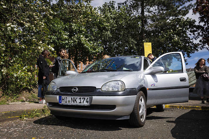 An outside view of a silver parked car that has the drivers side door open. People surround the car.