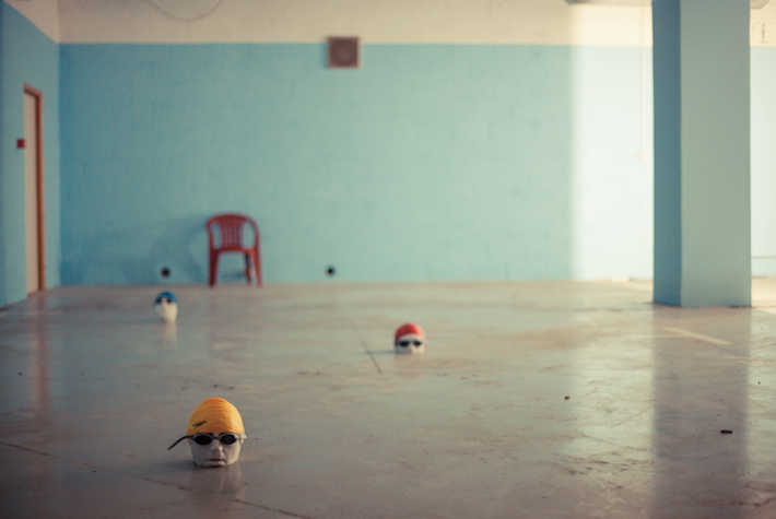 Le Swimming - sculptural heads in swimming goggles and caps laid out on the floor, as if in a swimming pool