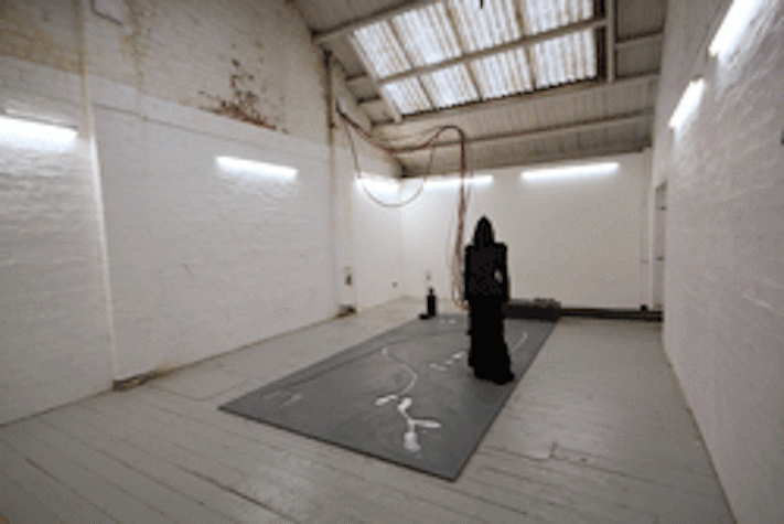 Gallery installation, sculpture of a clocked figure on a rectangular floor panel with shapes cut into it. A bundle of cables connect the figure to the wall. The gallery space has white walls, a pitched ceiling and a wooden floor