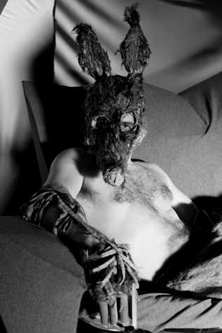 black and white film still. close up of topless man looking towards the camera. he wears a mask that resembles a rat, and a arm piece/glove