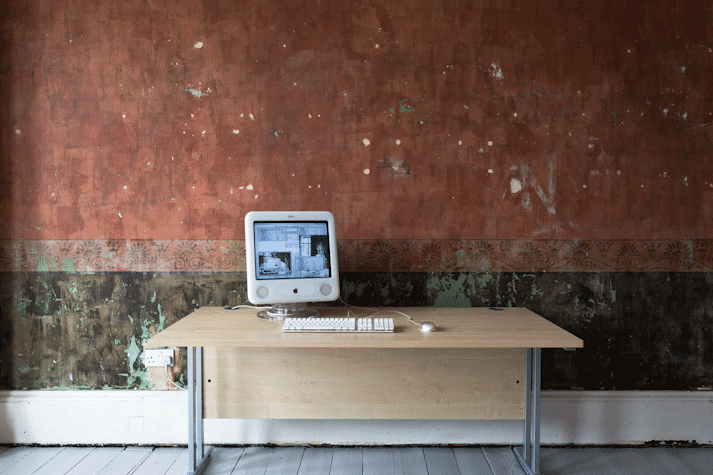 An early 2000s Mac computer sits on a beige laminate office desk with a keyboard and mouse. The screen has multiple different sized windows open, some with text some with glitchy black and white images.