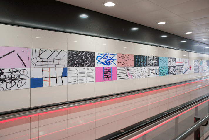 A close-up of a moving walkway with neon pink lights in Buchanan Street subway station. Two rows of colourful line-art artworks in the walls, fitted to the tiles.