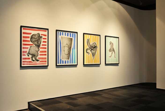 4 framed prints on a wall. the first has a red striped background and is a drawing of a monster/animal. the second has a blue striped background and is the image of a teapot tattoo. the third has a yellow striped background and a drawing of an elephant. the fourth has a green striped background and is a drawing of an anthropomorphic bird.