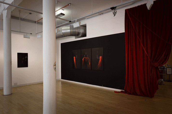 on a black wall at a distance three portrait photographs hang, showing a black figure on a black background holding a bright red drape of fabric