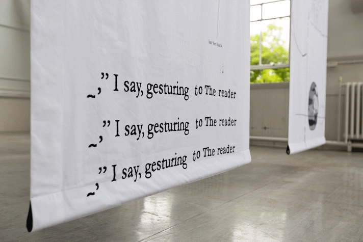 detail of bottom of white fabric hanging with black text that reads 'I say, gesturing to the reader' repeated three times over three lines