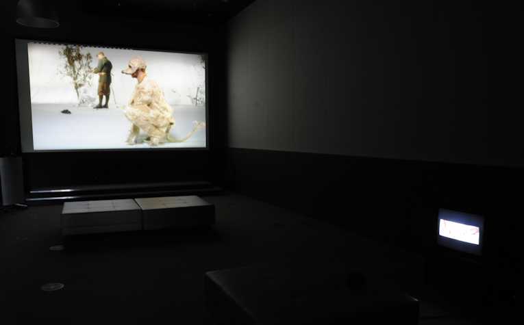 projection screen featuring a person dressed as a dog kneeling in front of a another person stood by a small tree