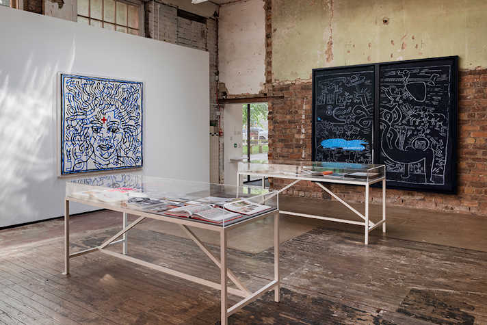 Artwork is displayed on brick walls in an industrial space. In the centre are are two glass-topped tables.