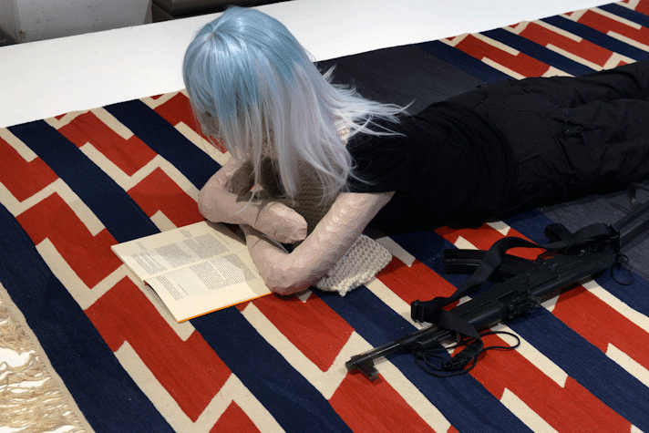 A manequin of a person with long blue hair with white wicks is reading a book lying down on a red, black and white rug. To their left is a gun. This is a part of an art installation in the Gallery of Modern Art, Glasgow International 2018.