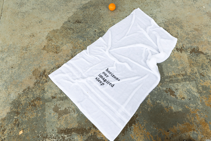 a white towel with black text that reads 'horizons our imagined sleep' is spread across a puddle of water on a gallery floor'