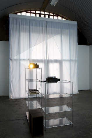 two long white curtains covering a window in a gallery space. in front are two metal and glass shelves with various objects and a projector