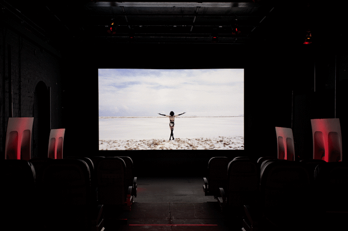 The image shows a projection screen. The screen shows a character stood with arms outstretched, back to the camera, in black lingerie and a black wig. They are in a barren snowy landscape.