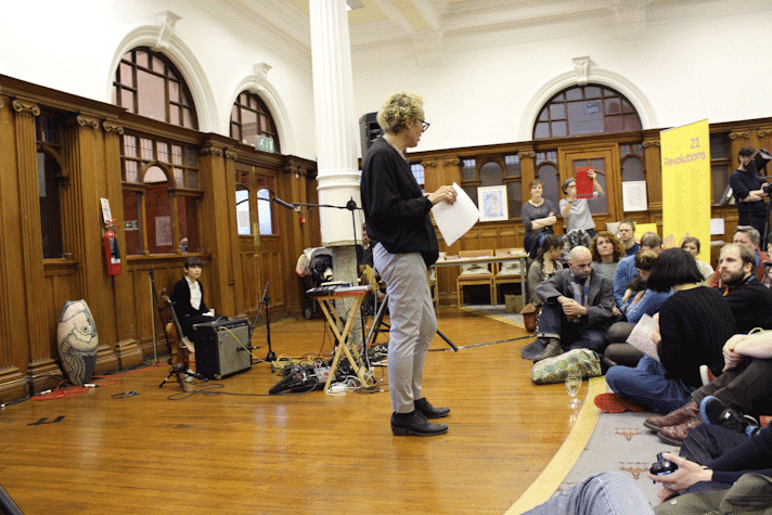 A woman is holding a piece of paper and facing a group of people sitting on the ground in front of her. The room is old fasg=hioned and wooden, with arches with glass over its doors.