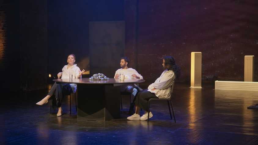 three people sat at a round table in Tramway 1 with mugs and glasses of water. behind them are three rectangular blocks, two vertical and one horizontal.