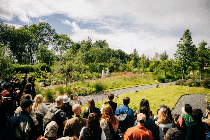 Audience members viewing a performance in a garden location with grass and flowers. Performers visible in the background