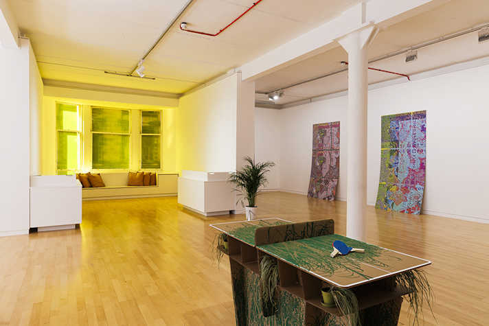 In a large white room, there is a ping pong table and prints displayed on the wall. At the far end of the room there is a sofa area and windows.