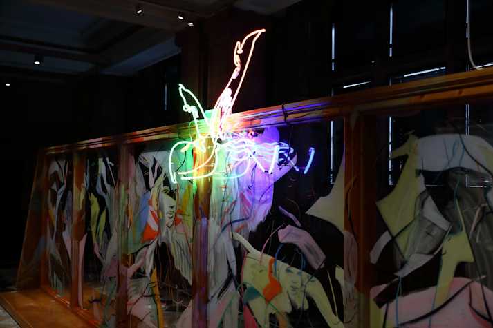 An abstract neon light showing two legs kicking the air in pointed heels, hangs at the top of a large wooden structure with glass panels and covered in fast gestural line paintings of figures and faces